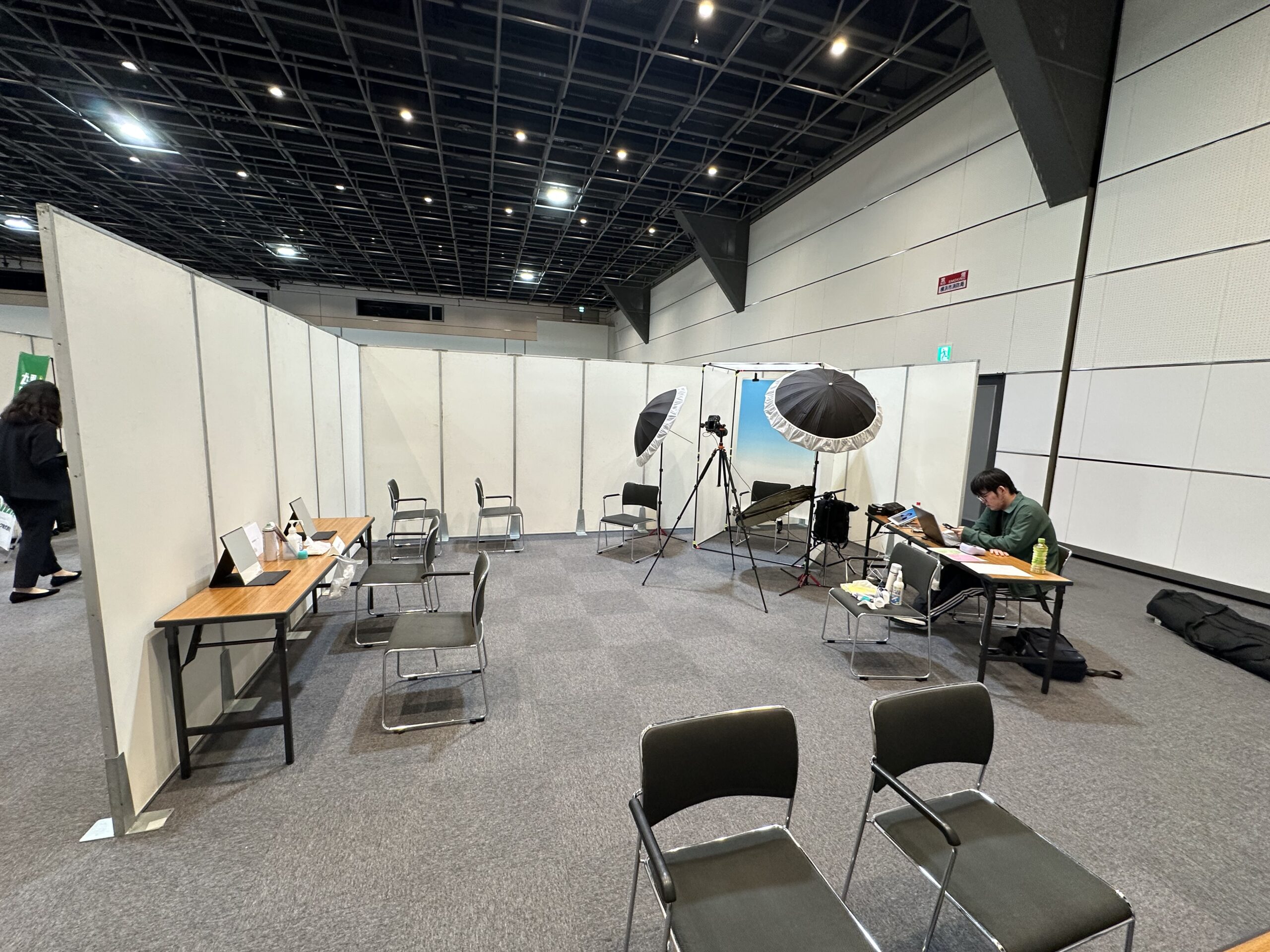 Indoor exhibition hall with white partition booths, empty chairs and tables, and photography lighting setup in the background.